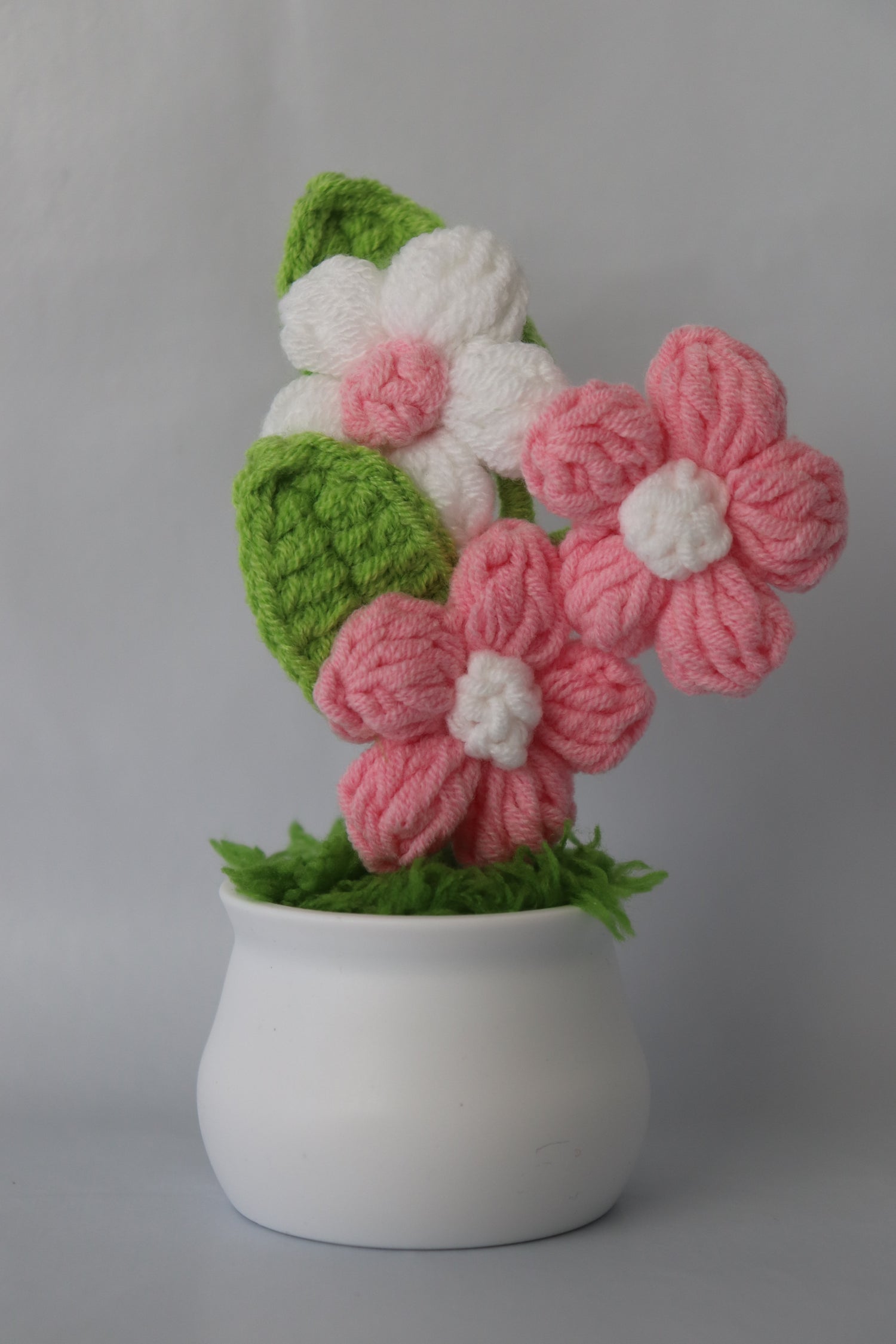 Knitted flowers in pink, white, and green on a white pot against a gray background