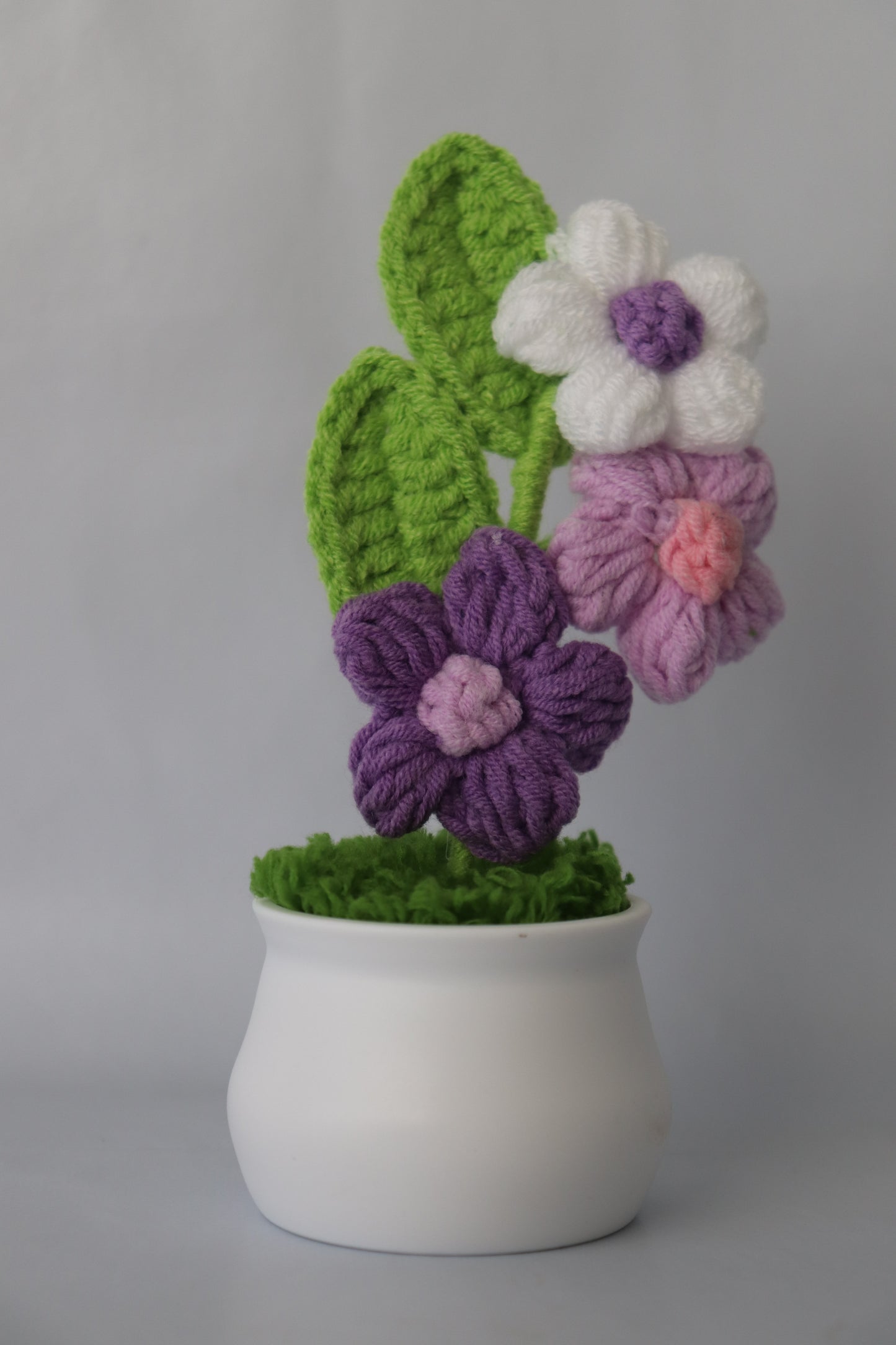 Crocheted flowers and leaves on a white vase against a gray background